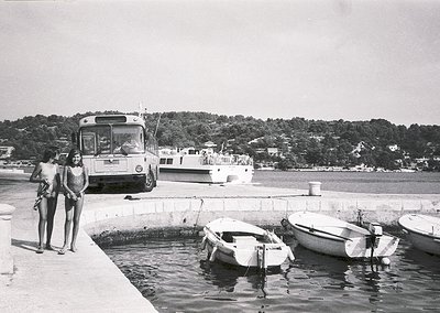 Two women in 1960s swimwear pose by a seaside pier, near a vintage ferry boat and small boats. Mid-century concrete harbor st...