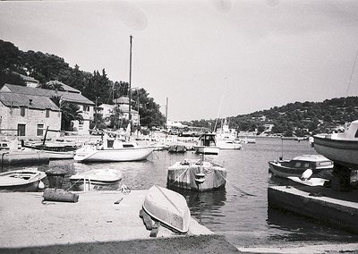 Black-and-white coastal marina with small boats docked along a concrete pier, surrounded by low-rise buildings with flat roof...