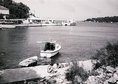 Mid-20th century seaside scene with a small motorboat docked near shore, two individuals sitting on rocky terrain. In backgro...