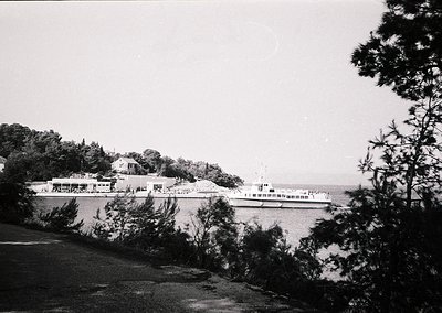 Black-and-white coastal scene featuring a mid-20th century ferry docked at a seaside promenade. Prominent low-rise buildings ...