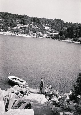 Black-and-white coastal scene featuring a man in mid-20th-century attire standing on rocky shore beside a small wooden boat. ...