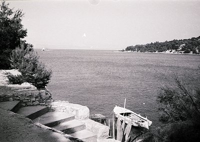 Black-and-white coastal scene featuring a stone staircase descending to a weathered wooden boat docked near calm waters. Dens...
