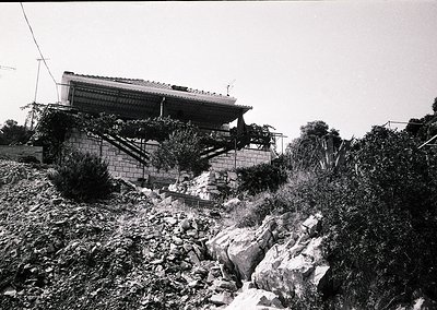 Damaged concrete structure perched on rocky terrain, likely post-earthquake or landslide. Overgrown vegetation and exposed fo...