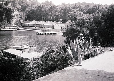 Mid-century lakeside resort with modernist pavilion and docked rowboat. Woman in striped swimsuit and hat stands near sun-ble...