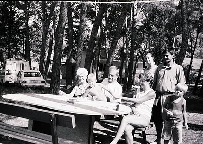 Family picnic in a wooded campsite, 1960s-70s. Six individuals seated/standing around a picnic table under tall trees; vintag...