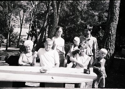 Family portrait in a shaded outdoor setting, likely mid-20th century. Six individuals—two adults, three children, and an infa...