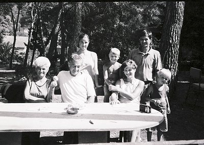 Family portrait in outdoor setting, likely mid-20th century. Six adults and three children pose around a picnic table under l...
