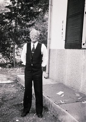 Mid-20th century man in vintage vest and tie poses outdoors beside a stone building. His posture suggests confidence; surroun...