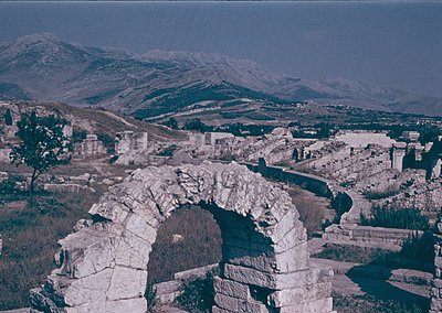 Ancient Roman triumphal arch fragment set against a mountainous landscape, likely part of a larger ruin complex. Mid-20th cen...