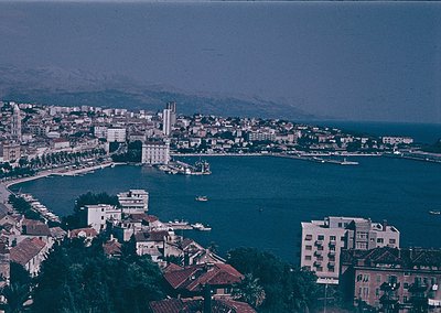 Aerial view of a coastal city with a prominent bay, featuring mid-20th century architecture. The scene includes a mix of low-...