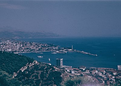 Aerial view of a coastal city with a prominent pier extending into the sea, flanked by mid-20th-century architecture. The tal...
