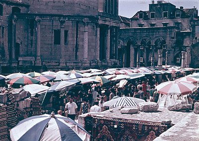 Vibrant 1960s/70s outdoor market in a historic European square, likely , Croatia. Roman-style arches and columns frame a bust...