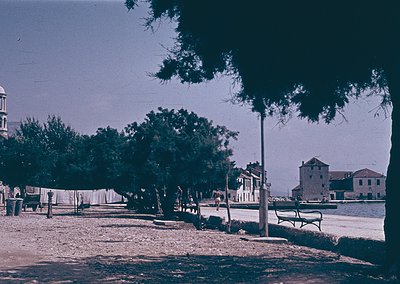 Vintage seaside promenade with mid-20th century architecture. A row of trees lines a paved walkway beside a body of water, wi...