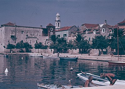 Vintage coastal scene featuring a historic stone fortress by the waterfront, likely Mediterranean. Two men in swim trunks wor...