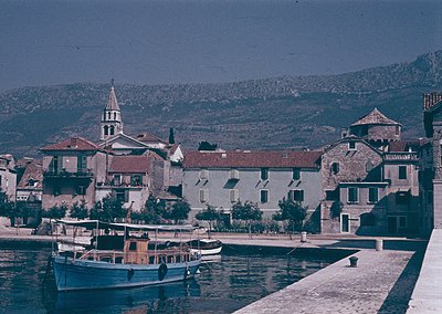 Vintage coastal town with Mediterranean architecture—stone buildings, terracotta roofs, and a prominent church steeple. Small...