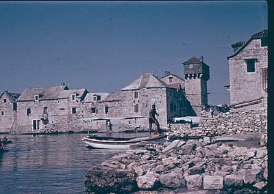 Historic stone fortress by waterfront, featuring medieval towers and low-rise buildings. A man in traditional attire stands n...