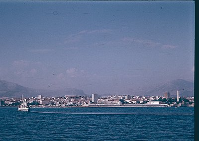 Vintage coastal cityscape with low-lying buildings, likely a Mediterranean port. A small boat floats in the foreground, while...