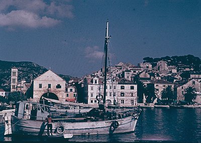 Vintage coastal scene featuring a small wooden fishing boat docked near a historic Mediterranean harbor. Whitewashed building...