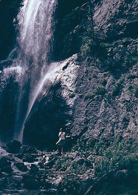 Black-and-white shot of a lone figure standing near a powerful waterfall cascading down rocky terrain. Dramatic lighting high...