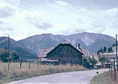 Vintage alpine village scene featuring a rustic timber-framed barn with steep gable roof, set against snow-capped mountains. ...