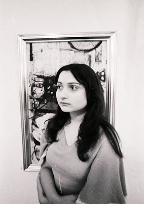 Portrait of a woman in a sleeveless dress, standing beside an abstract painting with bold, gestural brushstrokes and muted to...