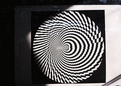 Optical illusion artwork featuring concentric, spiraling black-and-white stripes creating a dynamic vortex effect. Likely a 2...