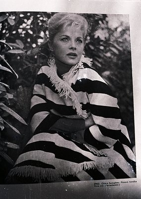 Vintage black-and-white portrait of a woman in a bold, striped sweater with a feathered collar, posed against a blurred green...