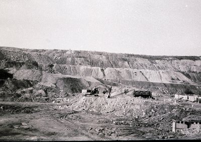Open-pit mining site with layered rock strata and heavy machinery—likely a bulldozer and conveyor system. Mid-20th century in...