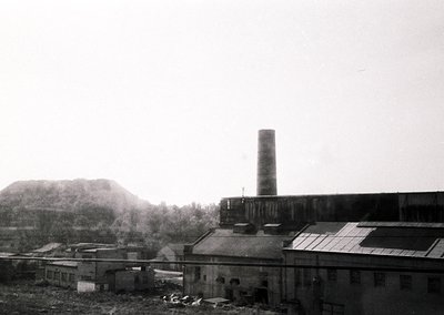 Industrial complex featuring a tall brick smokestack and multi-story factory buildings, likely mid-20th century. Overcast ski...