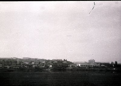 Vintage black-and-white coastal industrial scene featuring a long, low-lying building complex along a waterfront, likely a fa...