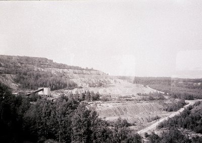 Mid-20th century industrial landscape featuring an open-pit mine with terraced excavation. Dense forest surrounds the site, c...