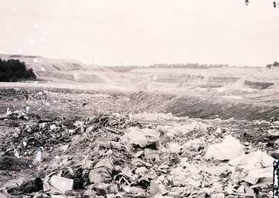 Mid-20th century industrial waste site with discarded materials including glass, metal, and debris. Open-pit excavation in ba...