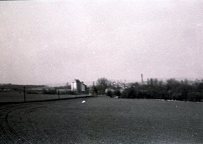 Mid-20th century industrial landscape with blurred motion, likely from handheld camera. Foreground shows a winding rural road...
