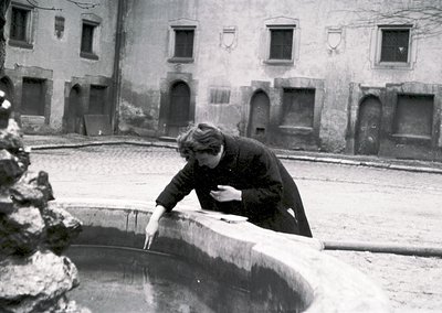 Black-and-white courtyard scene featuring a person in winter attire cleaning a stone fountain basin, surrounded by aged stone...