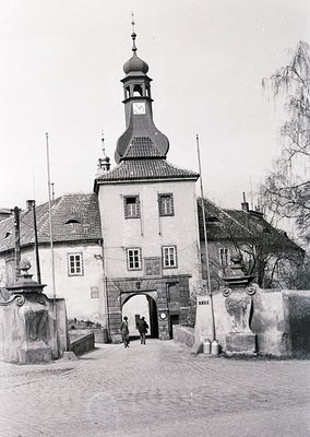 Historic town gate with Baroque-style tower and arched entrance, flanked by stone pillars. Cobblestone pathway leads through ...