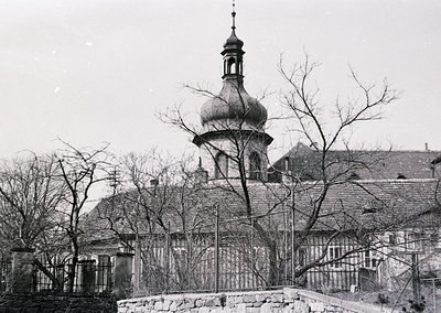 Black-and-white photograph of a domed church with a spire, framed by bare winter trees and a stone fence. Likely Eastern Euro...