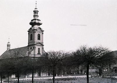 Neoclassical church with domed tower and symmetrical façade, flanked by trimmed trees in a courtyard. Mid-20th century Europe...