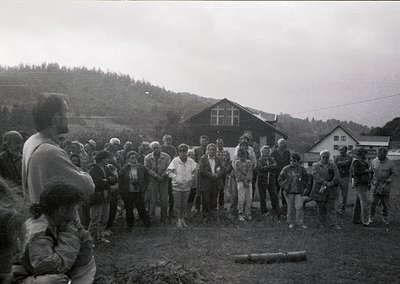 Black-and-white gathering of ~30 people in a rural outdoor setting, likely mid-20th century. Group stands on uneven terrain n...