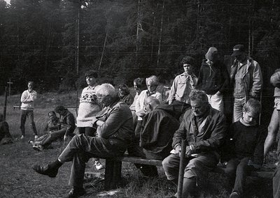 Group of 15+ individuals seated on wooden benches in a forested clearing, 1960s–1970s. Casual attire suggests a social gather...