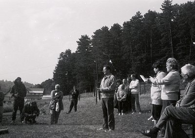 Black-and-white outdoor gathering in a rural setting, likely mid-20th century. A speaker addresses a seated audience of ~15 p...