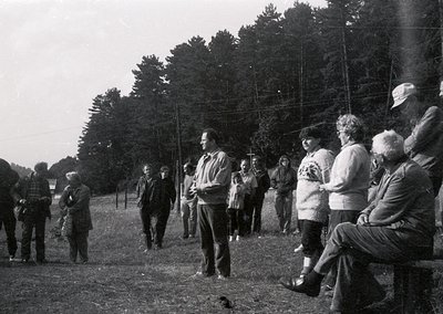 Group of adults in mid-20th-century outdoor gathering, likely 1950s–1960s. Central figure in dark jacket speaking to seated a...