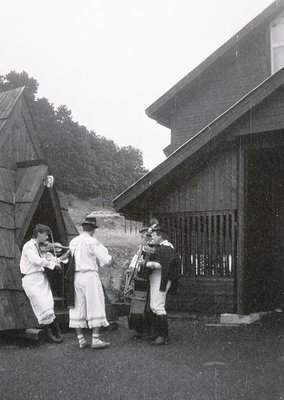 Three musicians in traditional folk attire play outdoors near rustic wooden structures, likely a rural setting. Violinist in ...