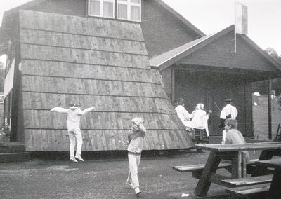 Black-and-white photo capturing a mid-20th-century outdoor scene. A man mid-jump against a wooden wall, while others observe ...
