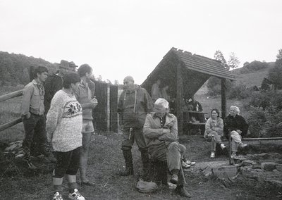 Group of 10+ individuals in a rustic outdoor setting, likely a hiking or camping area. Central figure seated on stone, surrou...