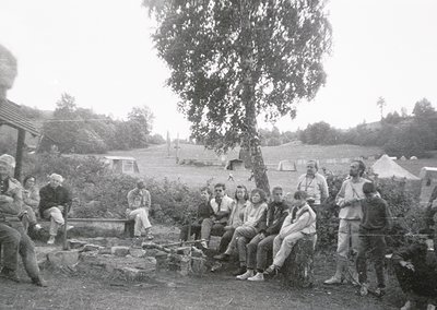 Group of 15+ individuals gathered around a campfire in a rural outdoor setting, likely 1960s–1970s. Wooden logs and makeshift...