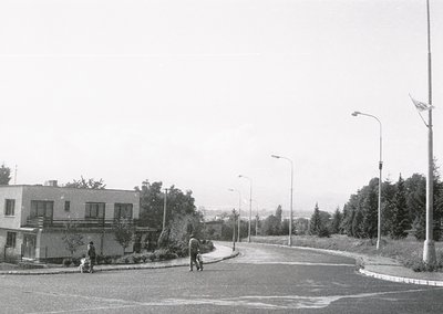 Mid-century urban street corner with minimalist Brutalist-style apartment blocks. Three pedestrians—one adult and two childre...