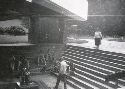 Vintage black-and-white photo of a grand, modernist stairwell with sweeping concrete steps. A man in mid-stride carries a lon...