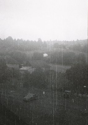 Black-and-white aerial view of rural landscape in heavy rain, featuring dense forest, open fields, and a lone tank-like vehic...