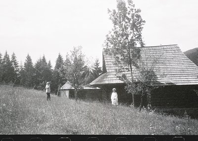 Black-and-white rural scene featuring a wooden house with shingled roof and stone foundation, surrounded by dense forest. Two...