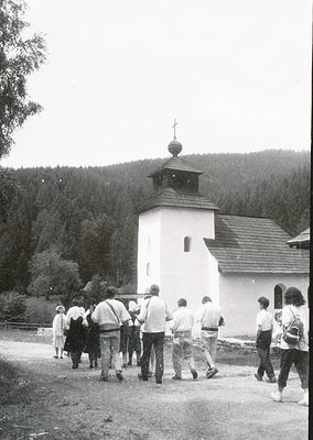 A group of people in casual 1970s attire—shorts, long-sleeve shirts, and backpacks—approaches a small, white Orthodox church ...
